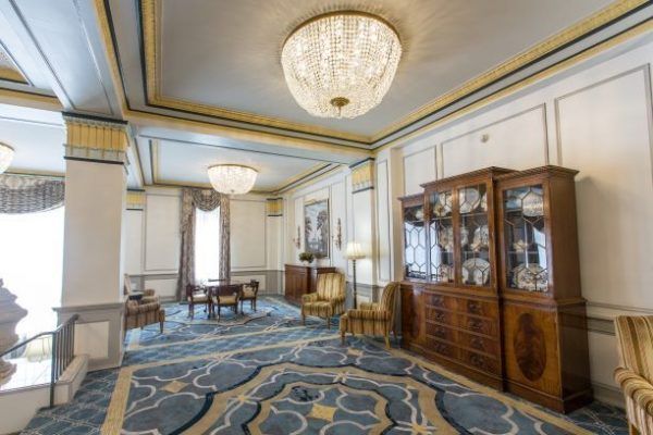 Lobby of the Francis Marion Hotel is shown with blues and yellows in the carpet along with antique wooden furnire and comfortable yellow Charis. Bright chandeliers shine from the ceiling.