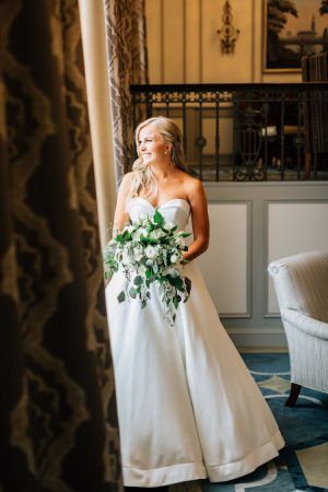 A bride in a white wedding dress that is floor length stands next a window. She is holding a bouquet of green and white flowers.