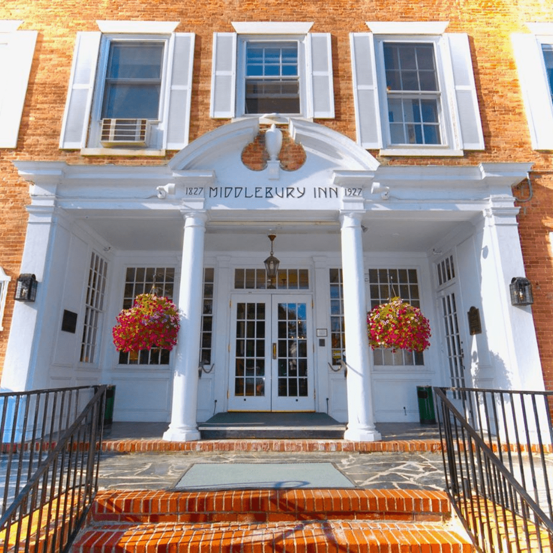 A brick building with white trim and two hanging baskets of flowers is shown. The Middlebury Inn reads across the op of the white molding of the entryway into the inn.