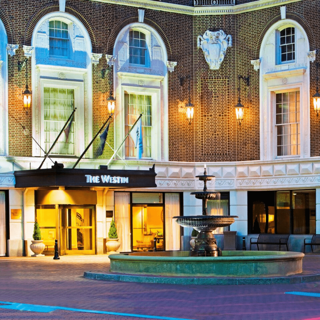 The façade of the Westin Poinsett hotel. A fountain sits in front of the doors and it is a grand, brick building with white molding and three flags fly above the westin signage.