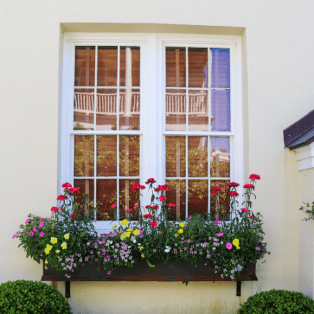 The edges of a yelloe house can be seen but the main focus is the window on a hsitpric home with a widnowbox full of beautiful, yellow, green pink and red flowers. the flowers vary in size, some tall and some are petite.